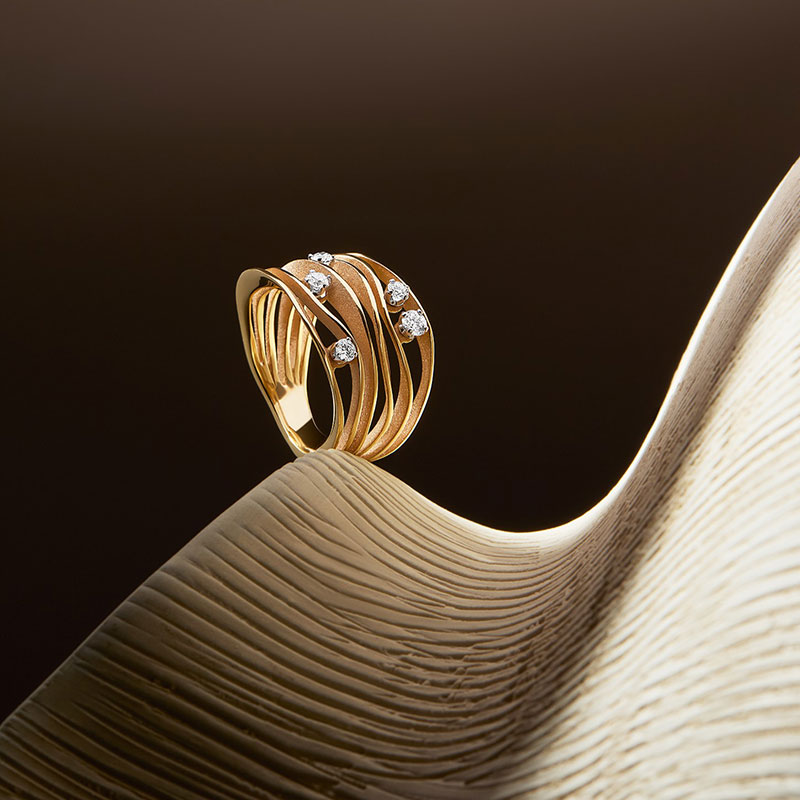 Ein goldener Ring mit funkelnden Diamanten liegt auf einer sandfarbenen gewellten Fläche