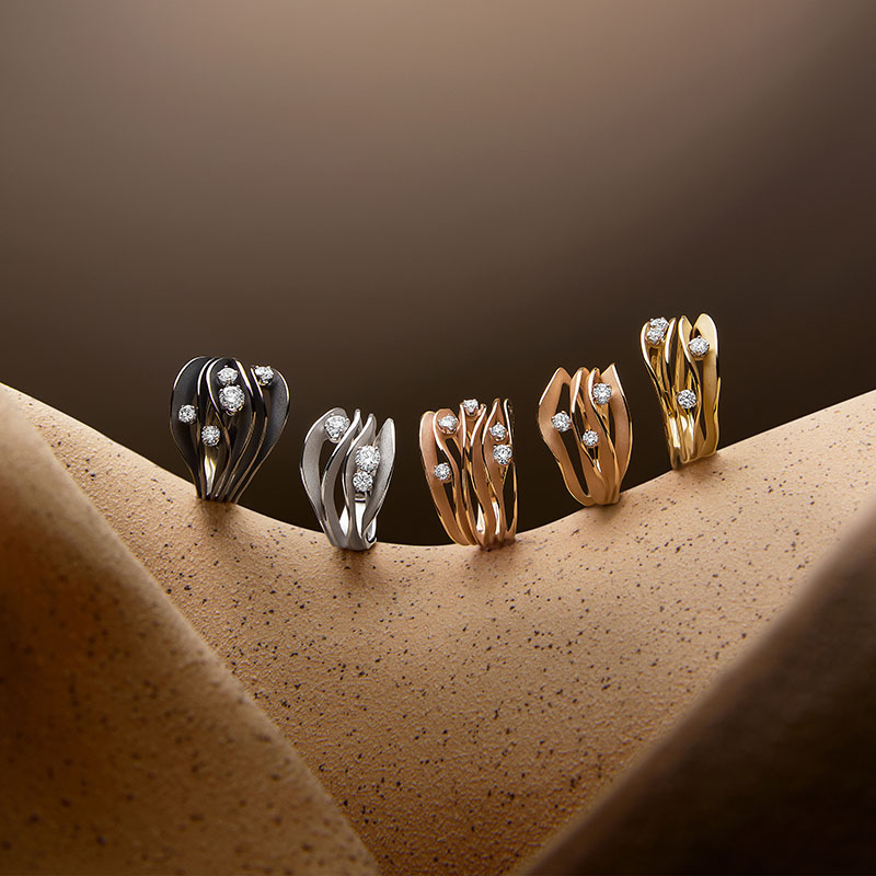 Fünf auffällige Ringe aus verschiedenen Metallfarben mit funkelnden Steinen auf sandfarbenem Untergrund