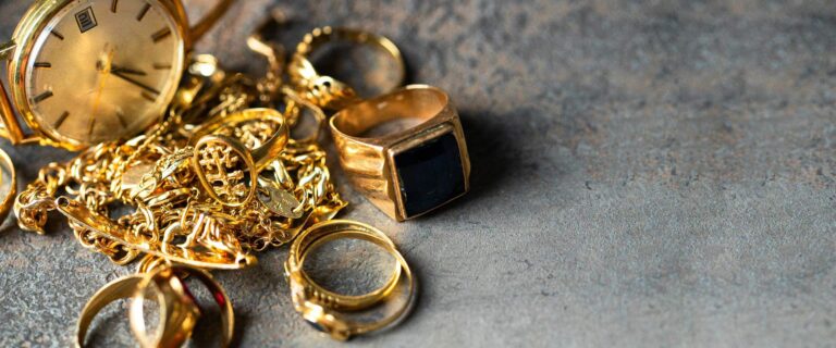 Mehrere goldene Schmuckstücke und eine Uhr liegen glänzend auf einem grauen Untergrund
