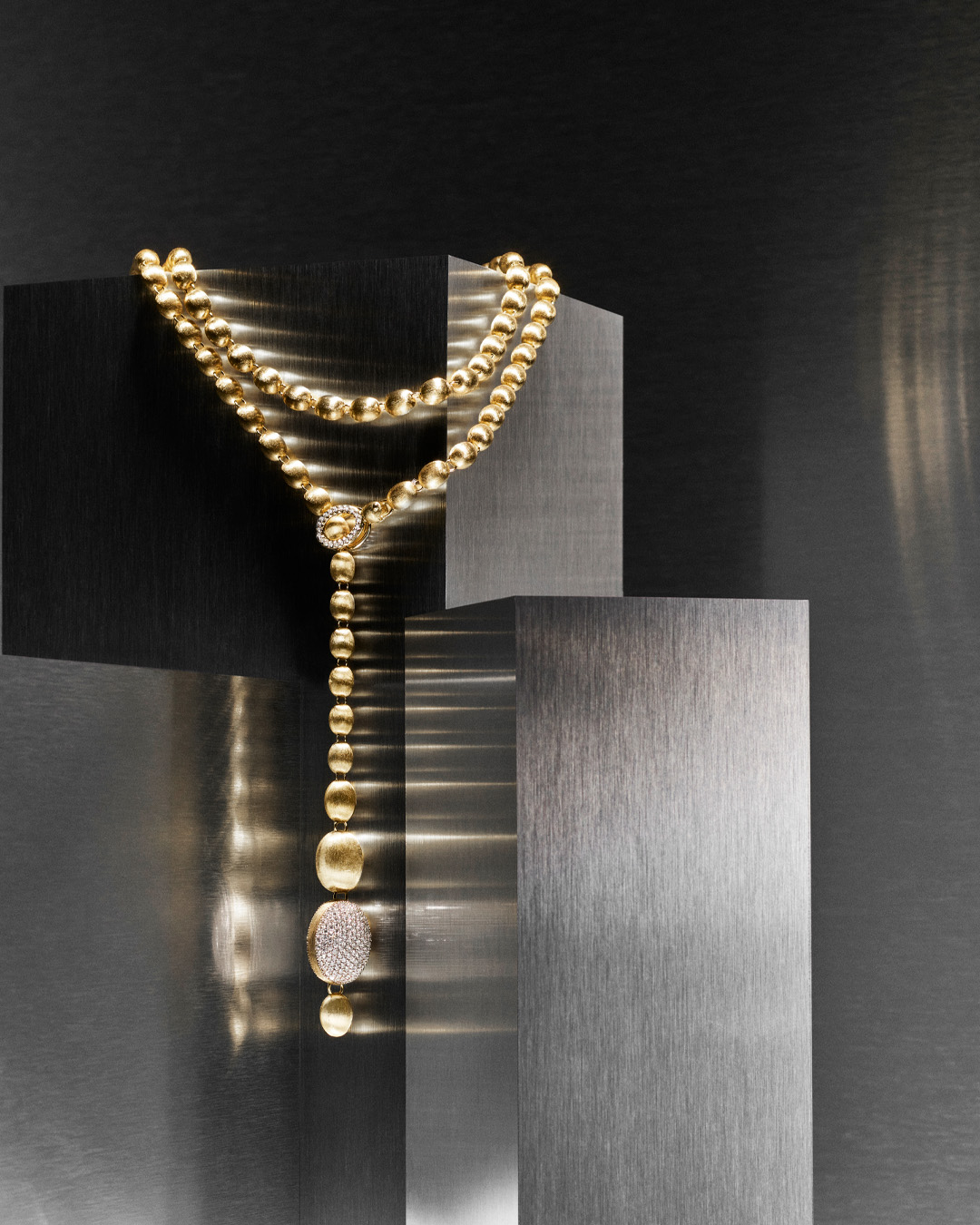 Eine goldene Halskette mit glänzenden Perlen liegt auf mattgrauen Metallblöcken