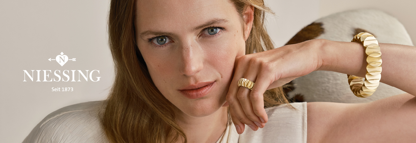 Junge Frau mit ernstem Blick trägt goldfarbenen Schmuck und helle Kleidung