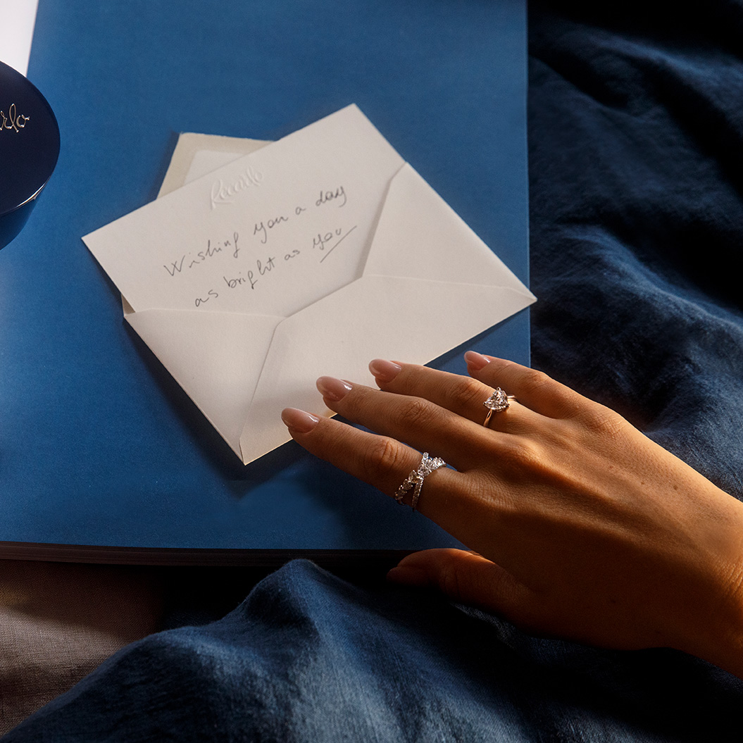 Eine Hand mit funkelnden Ringen öffnet einen weißen Briefumschlag auf blauem Stoff