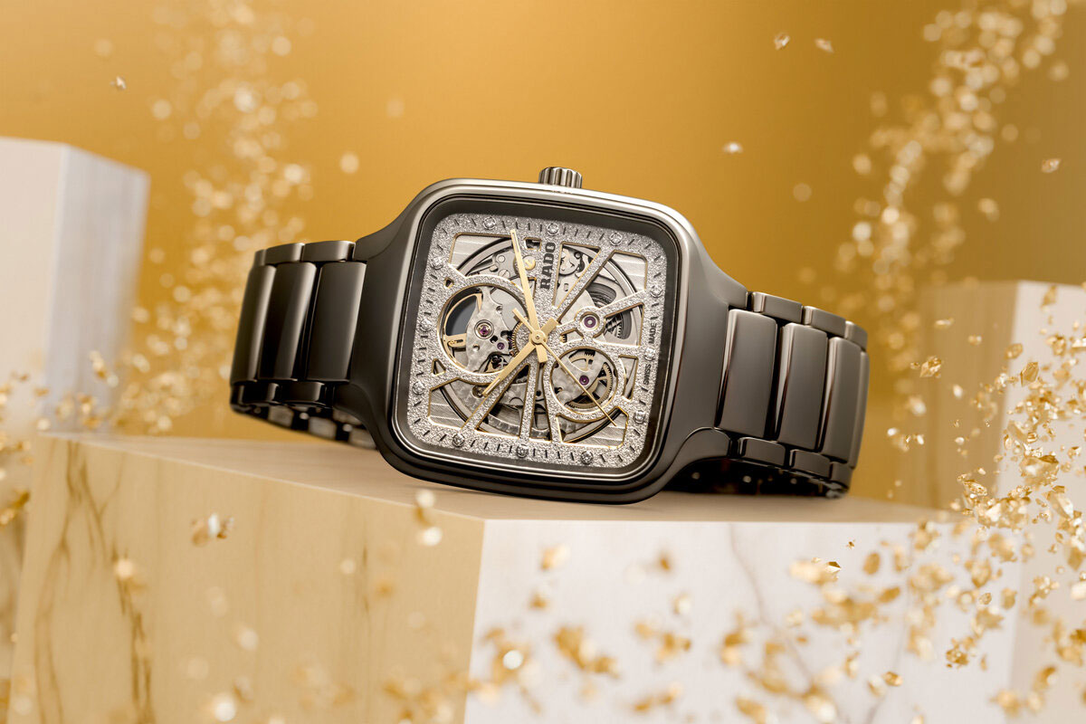 Schwarze Armbanduhr ohne Gesicht, glänzend, mit skelettiertem Zifferblatt in Gold und Silber auf hellem Podest vor goldenem Hintergrund mit Partikeln